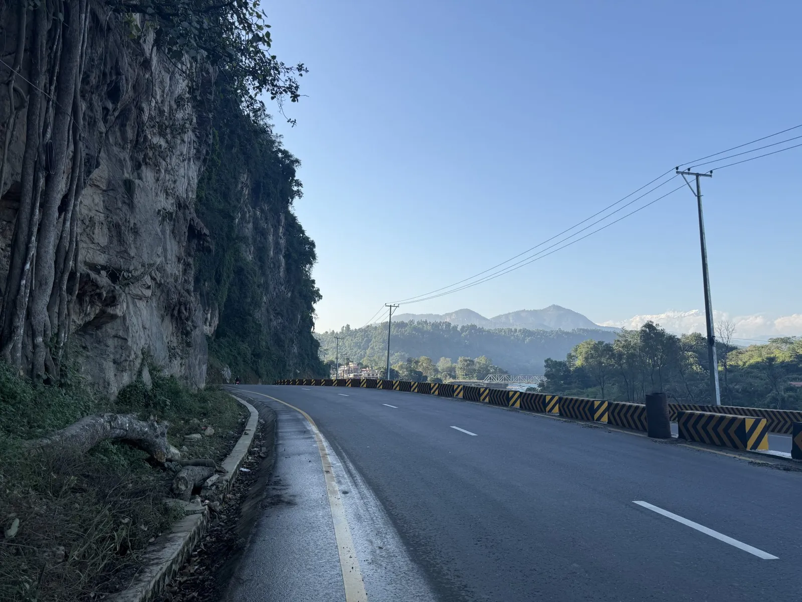 Estrada asfaltada em curva junto a uma parede de rocha coberta de vegetação, com montanhas ao fundo