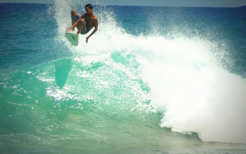 Surfista a fazer uma manobra no topo de uma onda com espuma branca e mar azul-turquesa