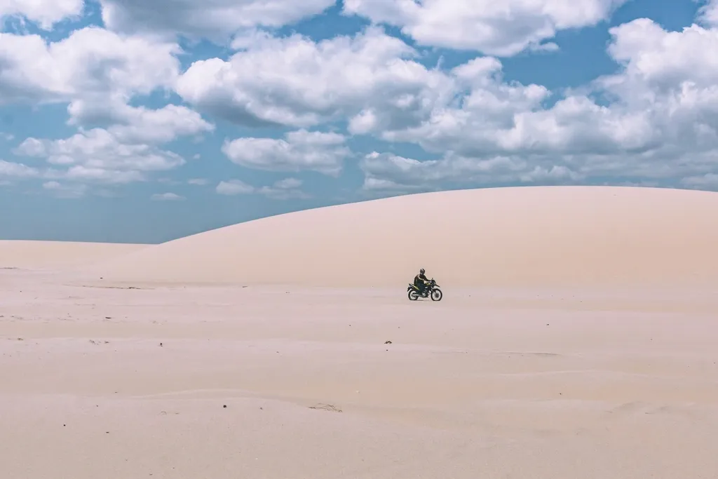 Motociclista isolado a atravessar uma duna de areia clara sob um céu de nuvens