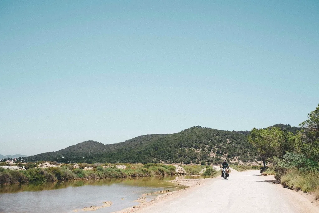 Motociclista numa estrada de terra junto a uma lagoa e colinas
